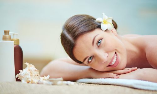 Happy girl with frangipani flower in hair looking at camera while relaxing in spa salon